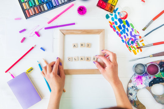 Top View Creative Art Class Concept. Female Hands Add Letter To Inscription On Canvas With Many Colorful Paintiing Materials On White Wooden Background. Drawing Lesson For Children And Adult. Flat Lay