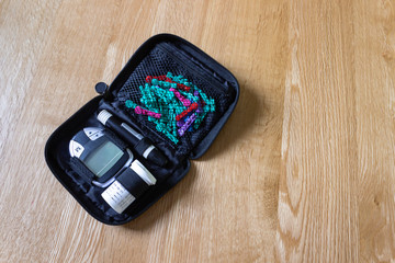 Diabetes blood glucose monitoring kit with electronic measuring device, container of testing strips and multi-color lancets in a black pouch on an oak surface