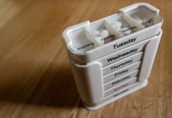 White seven day four compartment medication dispenser with medication standing on an oak surface. Subject lit with natural light from front