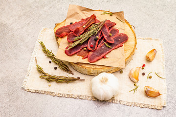 Basturma, dried tenderloin of beef meat, jerky, thinly sliced. Dry rosemary, pepper mix, chili, garlic on vintage linen cloth. Delicious food on a wooden board, stone background, close up.