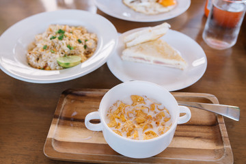 Breakfast table -set of cereal filled with milk in bowl and wooden plate, fried rice, Fried Egg, water and cream cheese sandwiches.