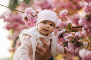 Cute baby girl between pink branches play with leaflets. Happy little girl surrounded by sakura