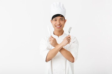 Image of asian positive chief man in white cook uniform smiling at camera while holding cutlery