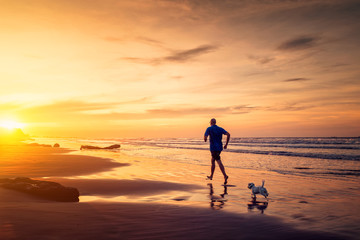 Man and small dog are running at the beach in sunset time