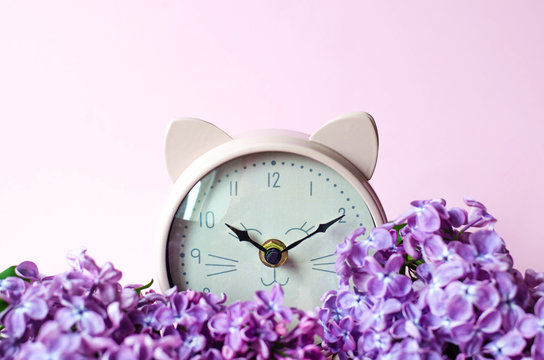 Pink alarm clock in the form of a cat lying on a bouquet of lilacs.
