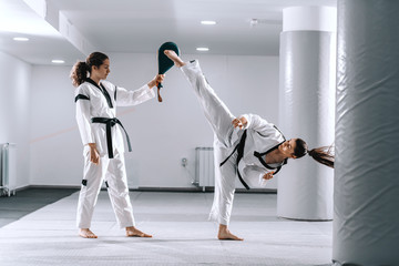 Beautiful Caucasian sporty girl in dobok kicking while other one holding kick target.