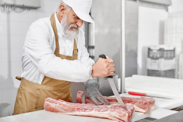 Man in uniform, apron cutting meat with knife on factory.