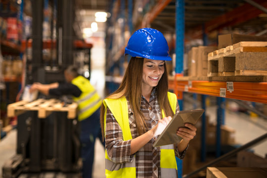 Female warehouse worker holding tablet checking inventory in distribution warehouse. Forklift in background.