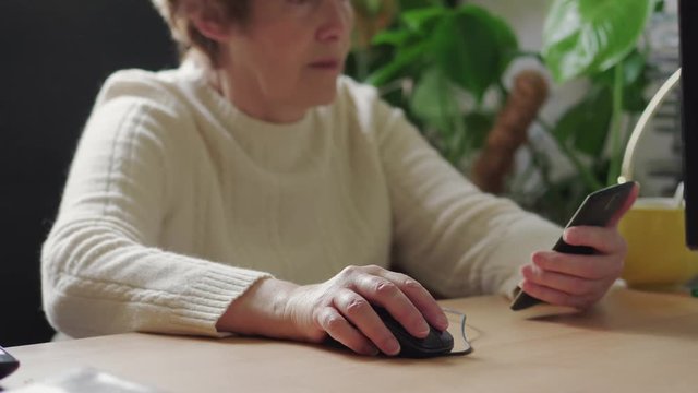 Senior woman clicking on mouse and keeping mobile phone in hand at work space. Elder businesswoman working with modern technology. Old lady browsing internet on laptop, taking cellphone. Grandmother