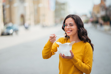 Millennial woman eating takeout food in the city street, smiling at camera.