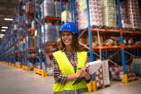 Portrait of female worker in distribution warehouse looking aside. Smiling woman with hardhat holding checklist.
