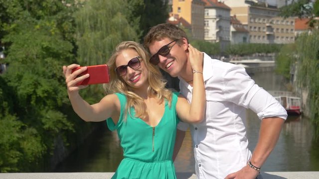 CLOSE UP, SLOW MOTION: Excited Caucasian man gives the thumbs up while taking a picture with girlfriend. Tourist couple video call home from Ljubljana while traveling around picturesque Slovenia.