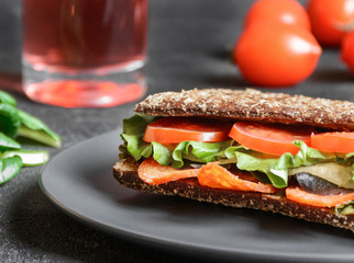 Panini - sandwich with brown bread, tomatoes, letuce, sausages, pieces of mozzarella and cup of coffee on dark background. Front view