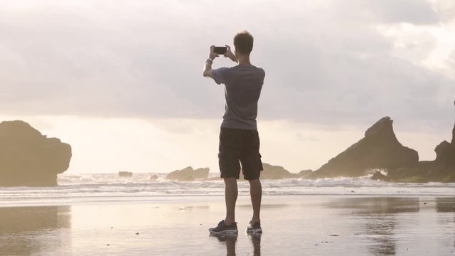 Man tourist taking pictures on smartphone camera on beach on the background of ocean and rocks. Bali, Indonesia