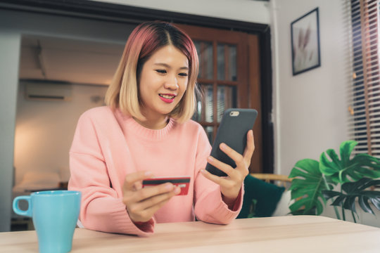 Beautiful Asian Woman Using Smartphone Buying Online Shopping By Credit Card While Wear Sweater Sitting On Desk In Living Room At Home. Lifestyle Woman At Home Concept.