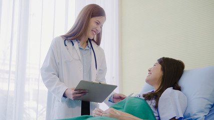 Beautiful smart Asian doctor and patient discussing and explaining something with clipboard in doctor hands while staying on Patient's bed at hospital. Medicine and health care concept.