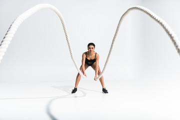 selective focus of sportive aafrican american girl doing exercise with ropes on white