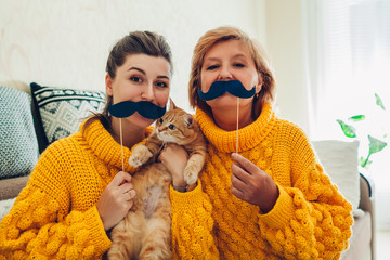 Senior mother and her adult daughter taking selfie with cat using photo booth props at home....