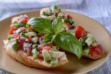 Two appetizing bruschettas with a garnish of finely chopped mix of various fresh vegetables, avocados, decorated with basil leaves on top, are on a plate, close up