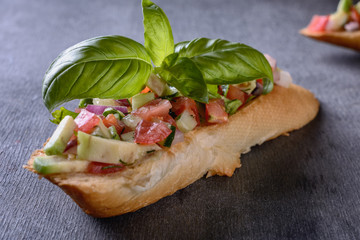 Appetizing bruschetta with a garnish of finely chopped mix of various fresh vegetables, avocados, decorated with basil leaves on top, are on a wooden dark board