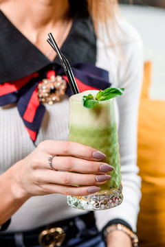 Young girl holding a glass with a green smoothie, decorated with basil, in a restaurant interior, sitting at the table. close up