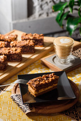 Chocolate brownie with nuts on a black plate on a yellow set on a wooden table in a restaurant. Cappuccino on the background. Close up. Space