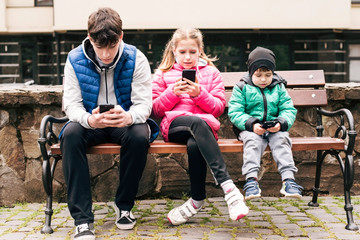 Three children sit on a bench and look at the phones, instead of walking, resting the children playing games on the phone