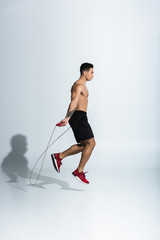 Fototapeta na wymiar side view of mixed race man in black shorts and red sneakers jumping with skipping rope on white