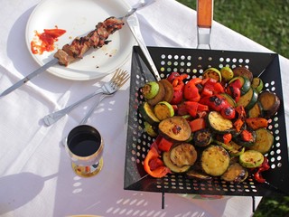 Frying pan with slice of vegetables, a plate of barbecue and a cup of wine on the white table outside