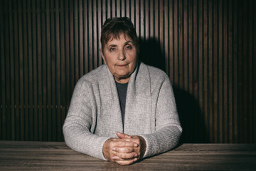 Portrait of poor senior woman sitting at table against wooden wall