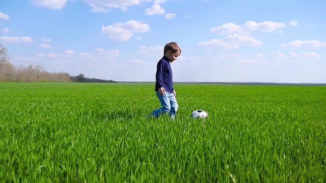 A happy child happily walks and kicks a soccer ball across a field of green grass in slow motion