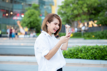 Young beautiful Asian lady celebrate with smart phone in the street in a sunny summer day, success happy pose, internet technology.