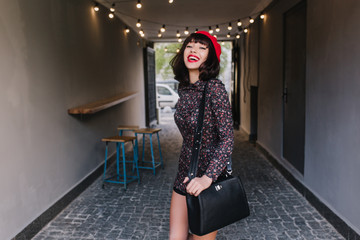 Gorgeous stylish girl with dark short hair walking after work carrying black leather bag and smiling. Portrait of adorable brunette young woman wearing vintage french clothes, having fun indoors