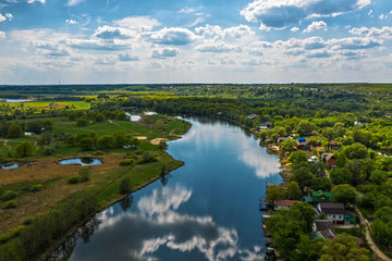 Aerial view of river with reflected blue sky and clouds, green meadows with trees and plants. Beautiful summer nature landscape from above