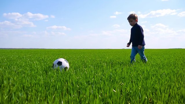 A happy child walks and kicks a soccer ball through a field of green grass in slow motion