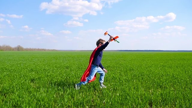 A little boy in a red cloak and mask holds an airplane in his hand and imagines flying