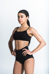 Fototapeta na wymiar beautiful sportive african american girl in black sports bra and shorts holding hands on hips on white background