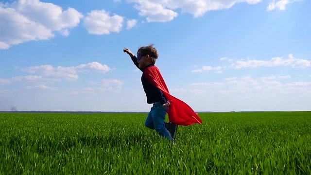 A happy child in a superhero costume in a red cloak runs across a green field