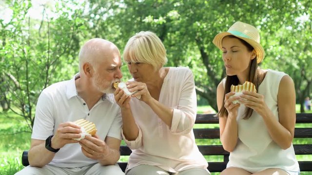 Senior couple with young teenage daughter sitting on bench and eating grilled sandwich with ham in public park. Hungry tourists family having lunch in garden enjoying summer sunny day