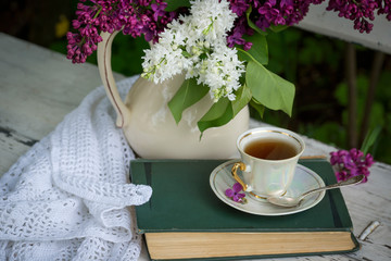 Beautiful branch of white and violet lilac flowers with opened violet note-book and black opened laptop, lying on the white wooden background with cup of tea, mock up perspective view