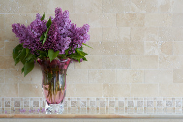 Bouquet of lilacs in glass vase on background wall of travertine. Flavor of spring flowers.