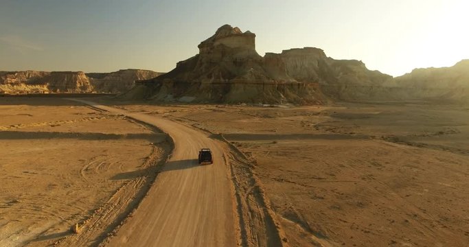 An aerial following long shot of a black 4WD car moving along the desert 4 An 4WD black offroad car moving along sandy road middle in great desert with mountains during golden hour long shadows