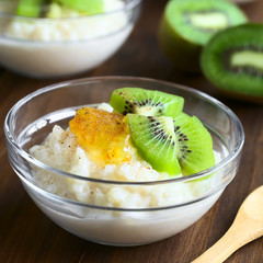 Rice pudding with kiwi pieces, orange jam and cinnamon in small glass bowl, photographed on dark wood with natural light (Selective Focus, Focus on the kiwi piece in the front)