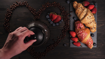 Obraz na płótnie Canvas On the background of a wooden table the heart of coffee beans in it a mug (coffee brewing) coffee can be seen steam. Next on the tray are croissants and fruit. Strawberries and blueberries around.