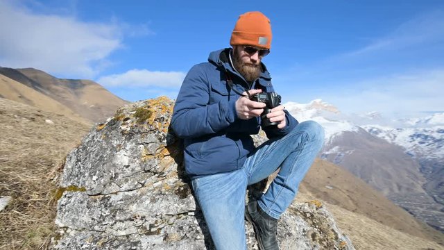 Portrait of a bearded traveler photographer in sunglasses and a cap sits on a rock with mirror camera in his hands and take a picture against the backdrop of mountains