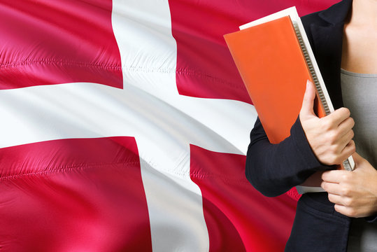 Learning Danish language concept. Young woman standing with the Denmark flag in the background. Teacher holding books, orange blank book cover.