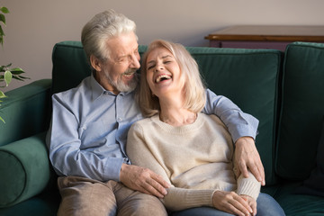 Loving middle aged husband embracing laughing wife, sitting on couch