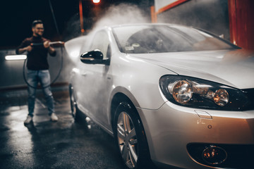 Young man washing his car in the evening at car wash station using high pressure water.