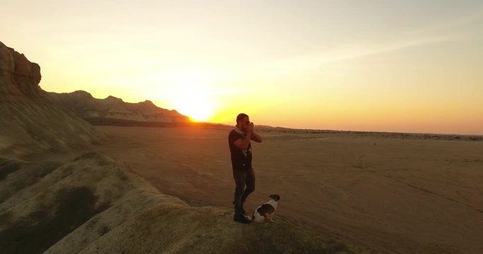 A rotating aerial shot of a man and his dog playing harmonica during sunset 3 An aerial shot of a man and his puppy standing on top of a hill  and playing harmonica long shadows golden hour