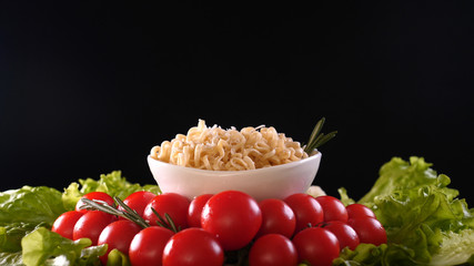 On a black background, vegetables and noodles on a wooden table, the whole dish is rubbed on top with parmesan cheese and mixed noodles. Concept of: Grater, Cheese, Noodles, Cherie Tomatoes, Salad.